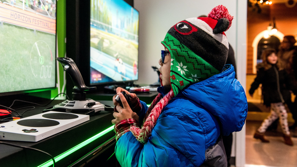 A child from the side view, seated and playing a video game on a console with a controller in hand. The individual is wearing a colorful beanie with a pom-pom and a blue jacket. In front of the person is a screen displaying what appears to be an in-game car racing scenario. The setting seems to be indoors, possibly at an electronics store or gaming area, as indicated by the ambient lighting and other items in the background that are not fully visible due to blurring. There is also green lighting emanating from beneath the screen, which adds to the gaming atmosphere. A portion of the individual’s face has been obscured with a green block for privacy reasons.